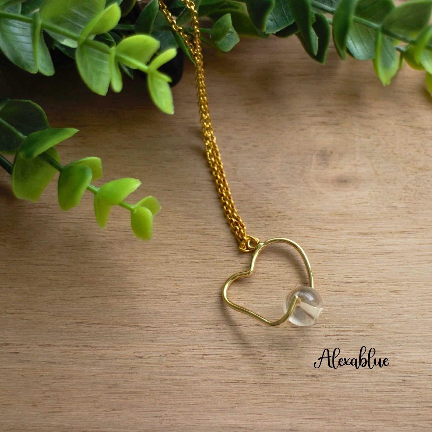 Gold chain necklace with heart-shaped wire pendant and clear stone on wood, greenery nearby