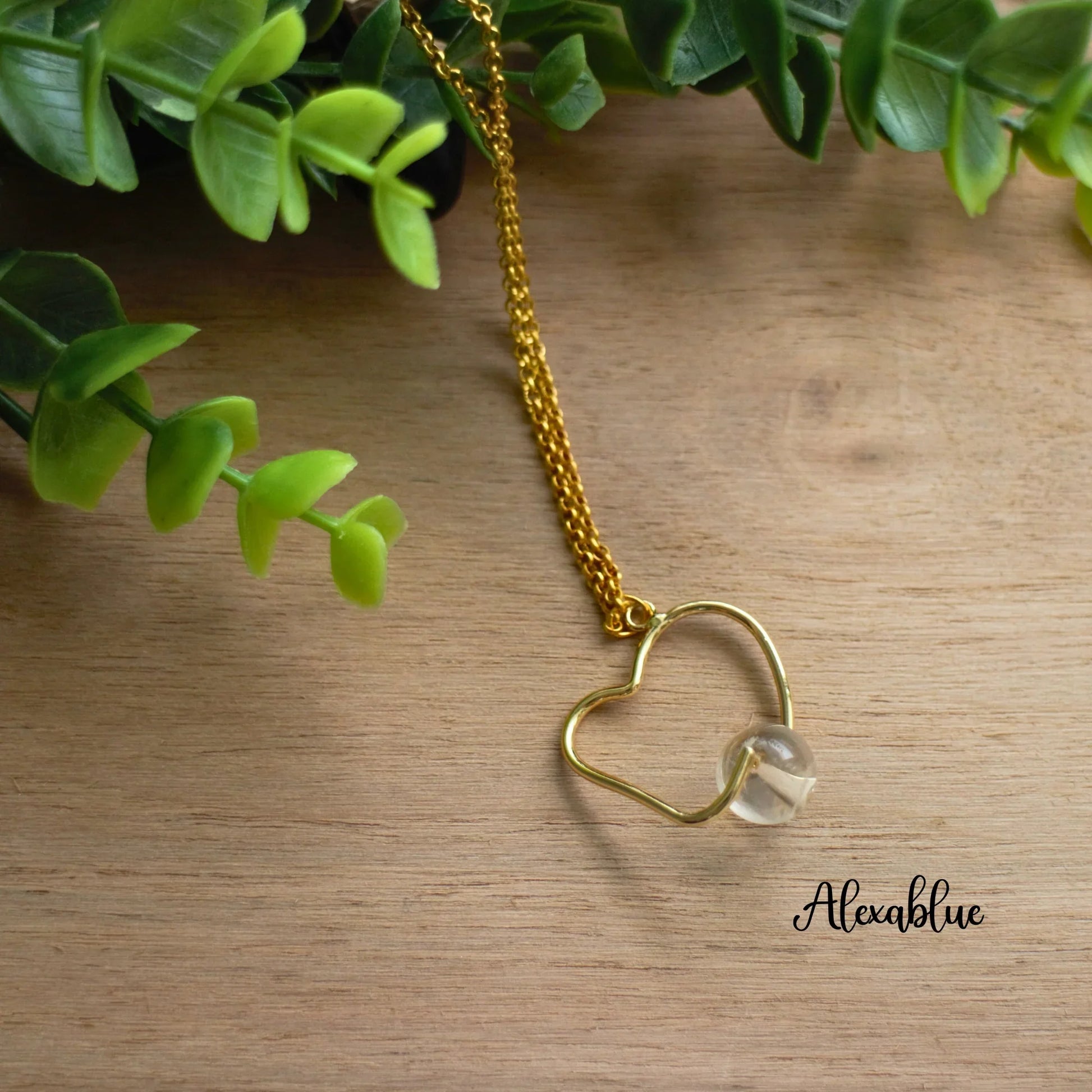 Gold chain necklace with heart-shaped wire pendant and clear stone on wood, greenery nearby