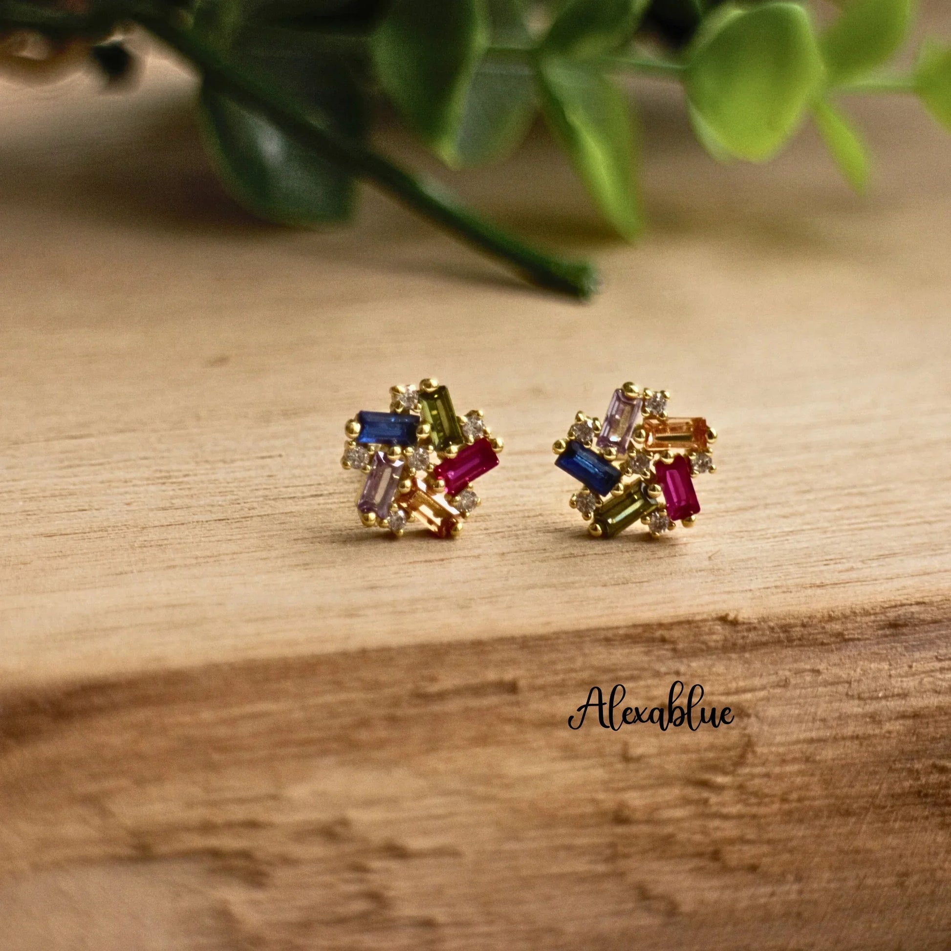Gold stud earrings with multicolored rectangular stones on wooden surface, plant background
