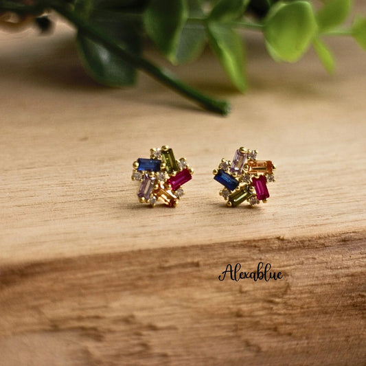 Gold stud earrings with multicolored rectangular stones on wooden surface, plant background