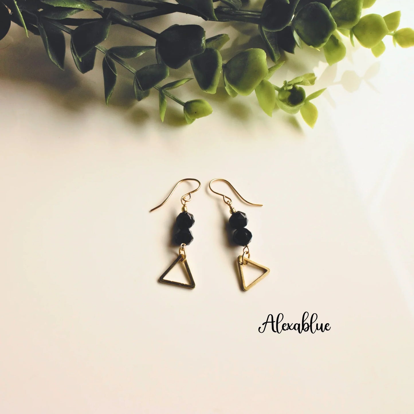 Gold geometric dangle earrings with black natural stones on a white background and green leaves.