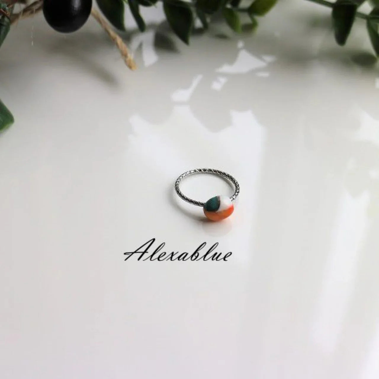 Silver twisted band ring with multicolor stone bead on white surface, surrounded by greenery.