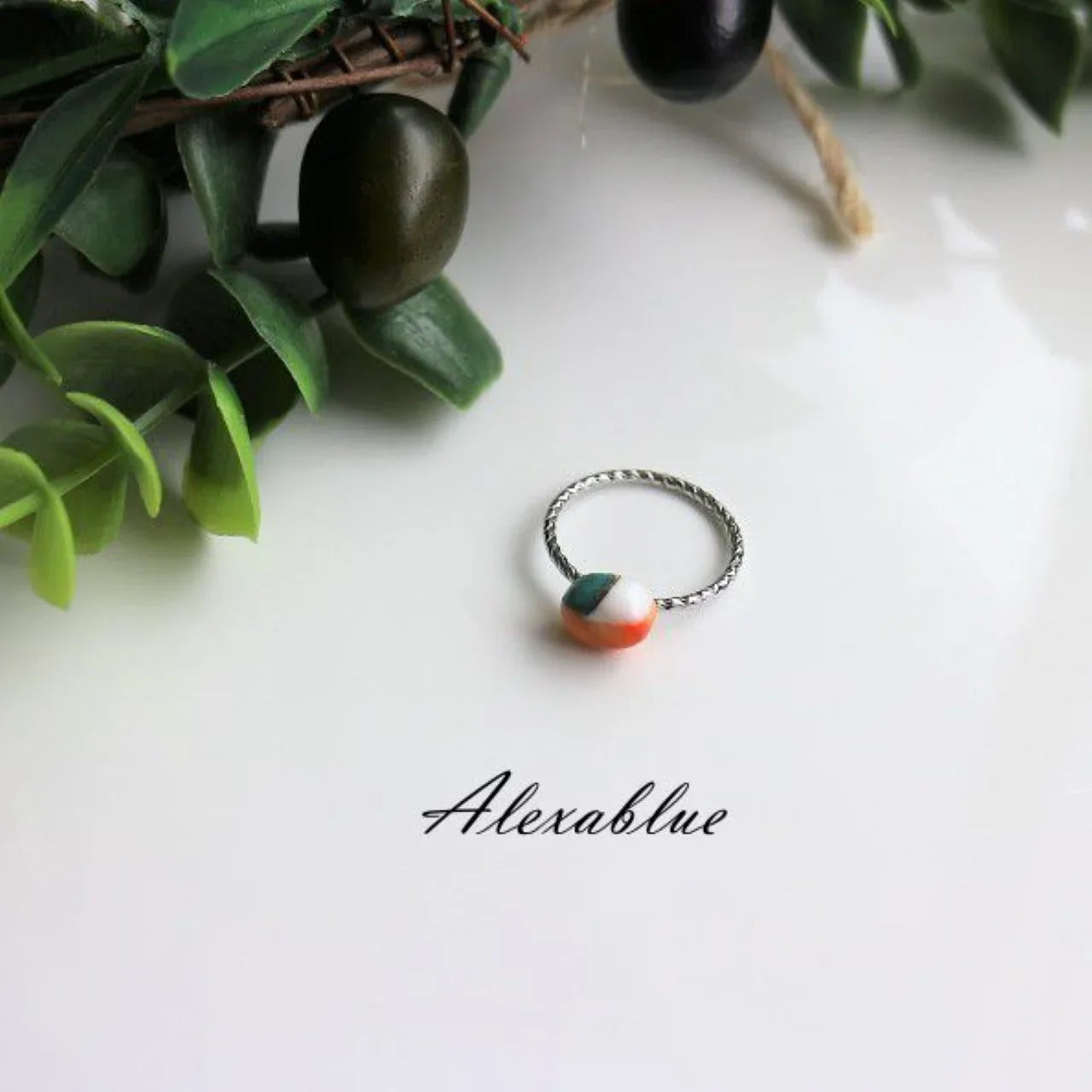 Silver twisted band ring with a round multicolored stone, displayed on a white surface with green leaves.
