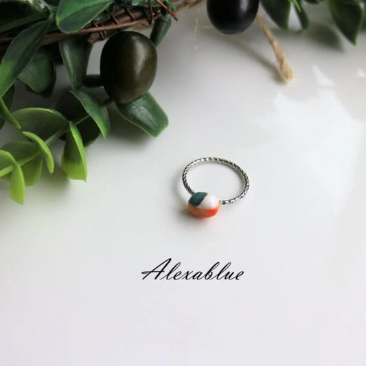 Silver twisted band ring with a round multicolored stone, displayed on a white surface with green leaves.